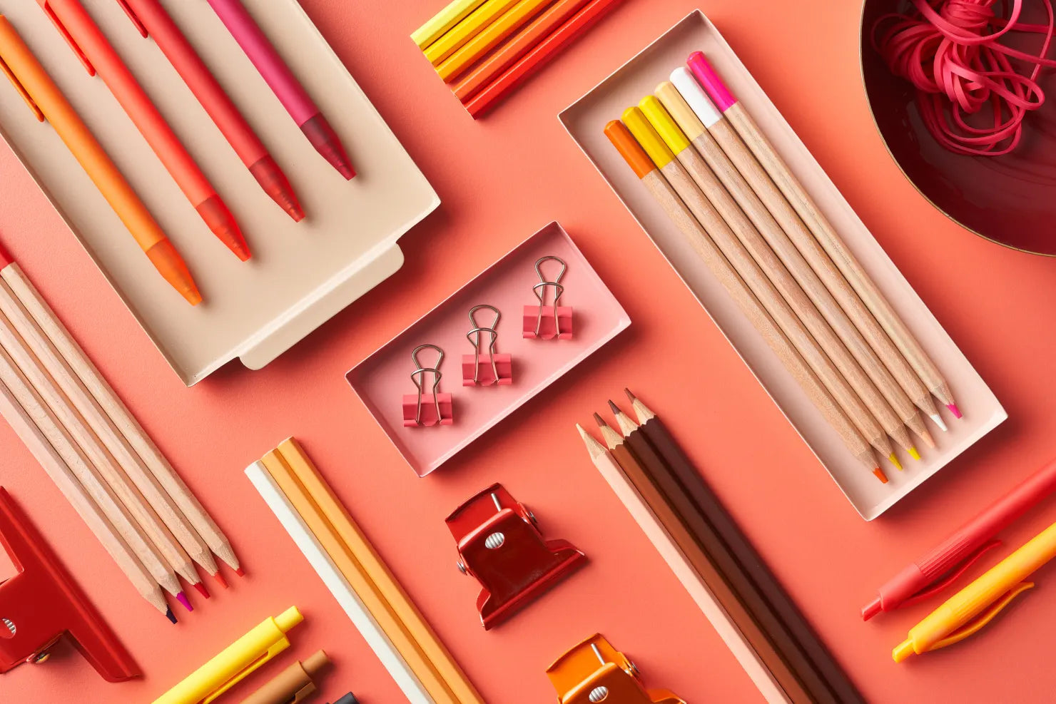 accessoires de papeterie avec crayons, stylos, pinces de photographe de couleurs orangés à rose posés de manière organisés sur une table de couleur saumon